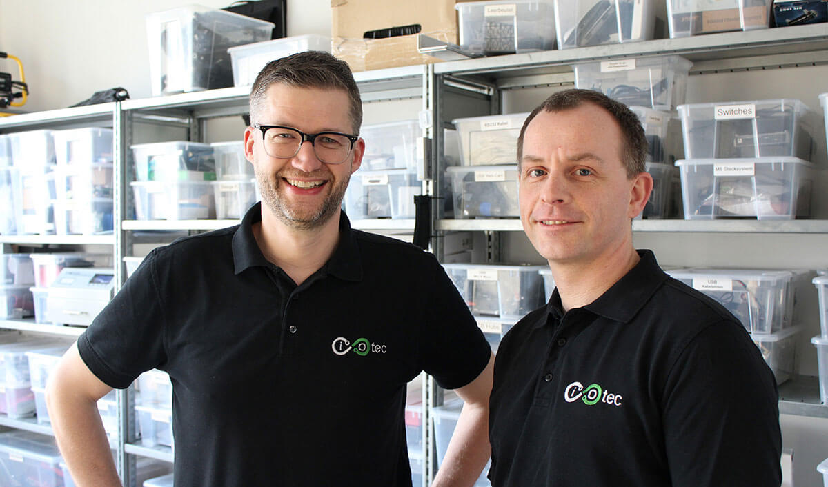 Zwei lächelnde Männer in schwarzen Poloshirts mit iotec-Logo stehen vor Regalen mit beschrifteten Sortierboxen.