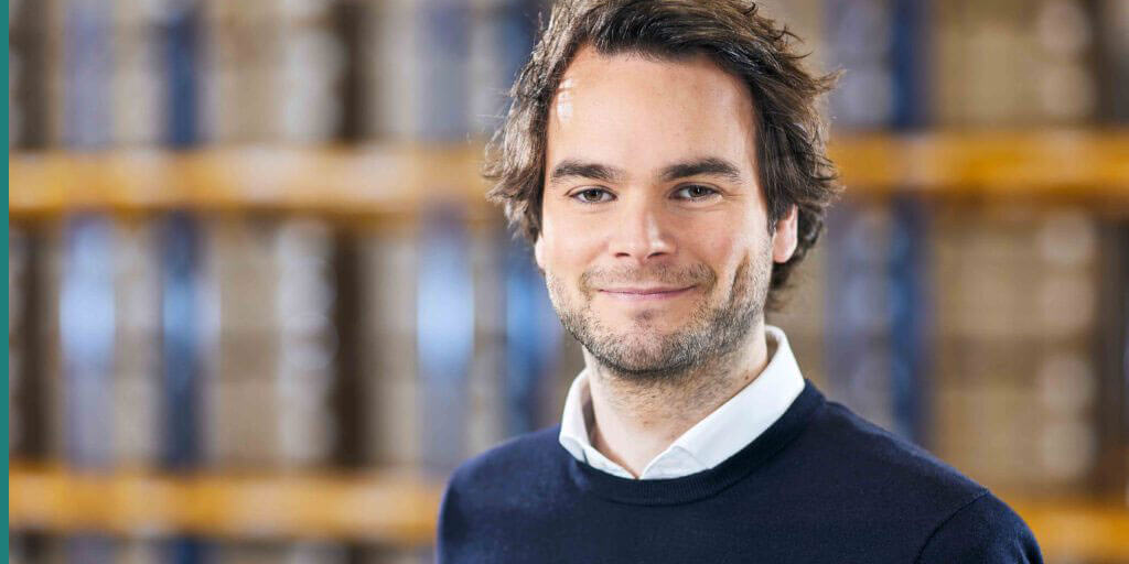Portraitfoto von Jan Steinacker, lächelnd, vor einem unscharfen Regal im Hintergrund.