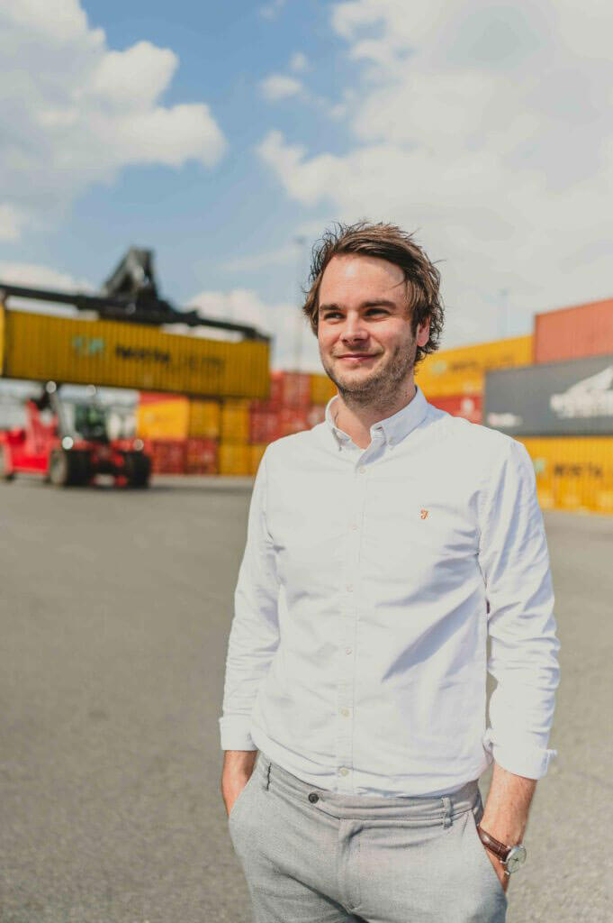 Portraitfoto von Jan Steinacker, lächelnd, in einem Hafen mit Containern und einem Gabelstapler im Hintergrund.