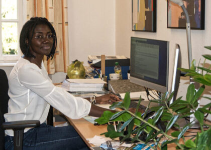 Joyce Noufele sitzt lächelnd an einem Schreibtisch vor einem Computerbildschirm im Büro.