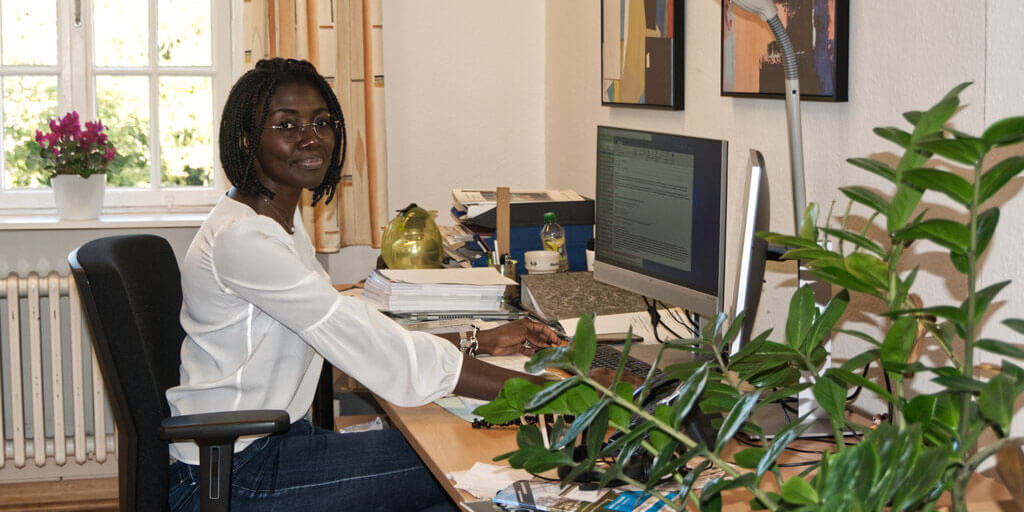 Joyce Noufele sitzt lächelnd an einem Schreibtisch vor einem Computerbildschirm im Büro.