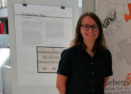 Portraitfoto von Judith Franzen, lächelnd, vor Informationstafeln zu einer Ausstellung über den Dreißigjährigen Krieg.