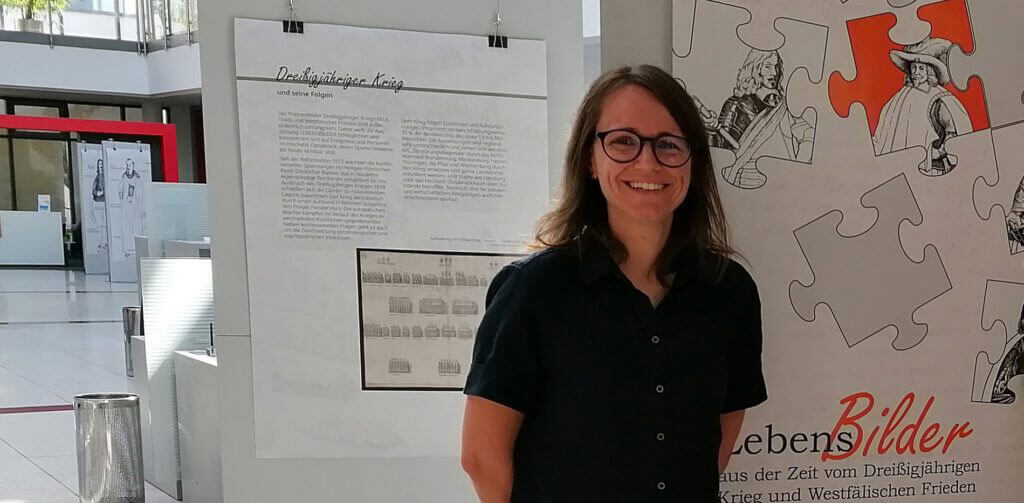 Portraitfoto von Judith Franzen, lächelnd, vor Informationstafeln zu einer Ausstellung über den Dreißigjährigen Krieg.