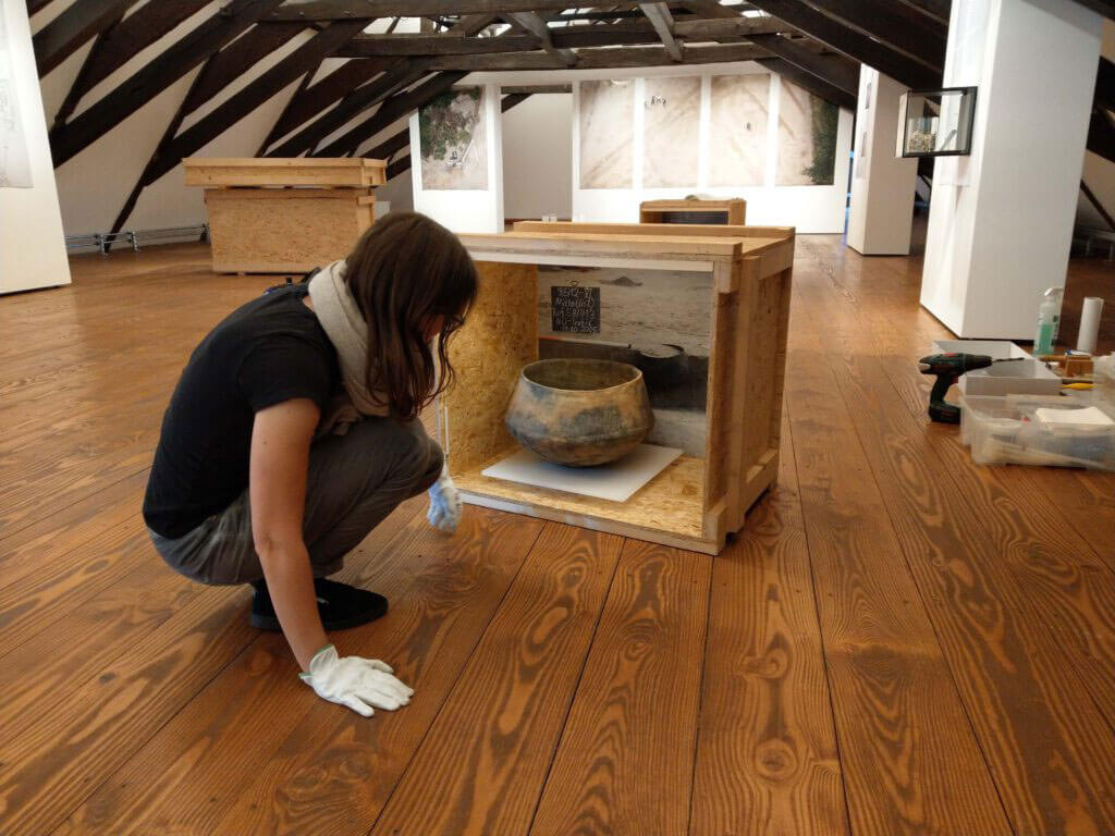 Judith Franzen hockt in einer Ausstellungshalle und betrachtet eine archäologische Tonscherbe in einer Holzkiste.