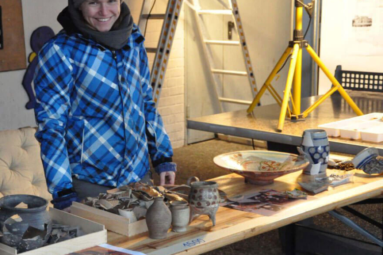 Judith Franzen steht an einem Tisch mit archäologischen Fundstücken, darunter Keramikgefäße, und lächelt.