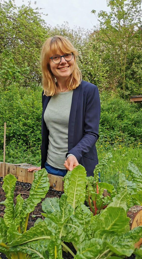 Julia Fink lächelt und steht in einem Garten hinter einem Hochbeet mit grünem Mangold.