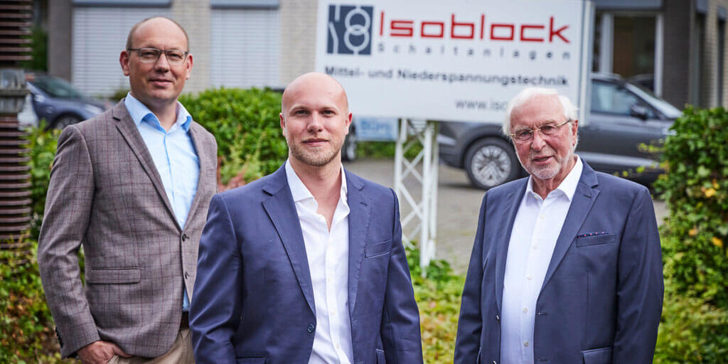Drei Männer im Anzug bzw. Sakko stehen nebeneinander vor einem Firmenschild von Isoblock Schaltanlagen.