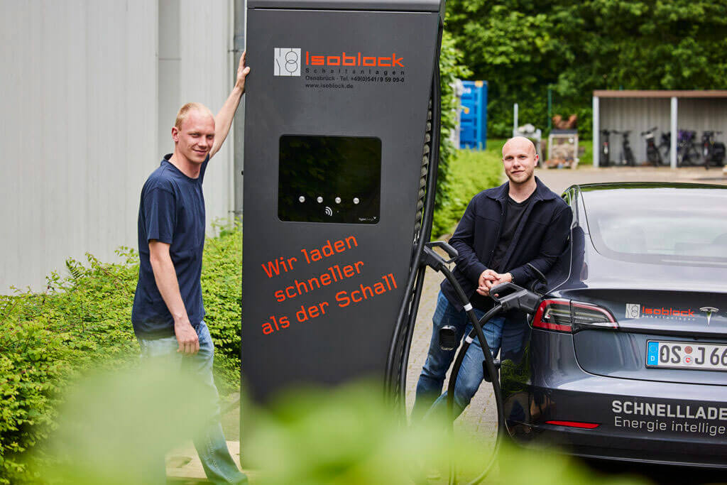 Zwei Männer stehen lächelnd an einer Isoblock-Schnellladestation neben einem Tesla-Elektroauto.