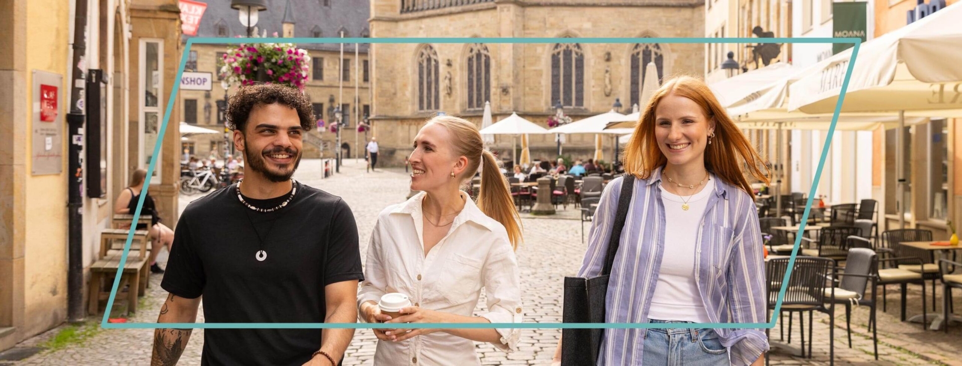 Drei junge Menschen gehen lachend über einen gepflasterten Platz in einer historischen Altstadt.