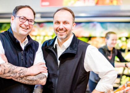 Jörg Kaluscha und Oliver Wehling lächeln nebeneinander in der Obst- und Gemüseabteilung eines Supermarkts.