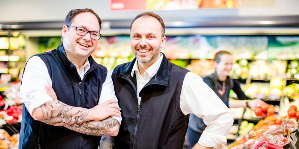 Jörg Kaluscha und Oliver Wehling lächeln nebeneinander in der Obst- und Gemüseabteilung eines Supermarkts.