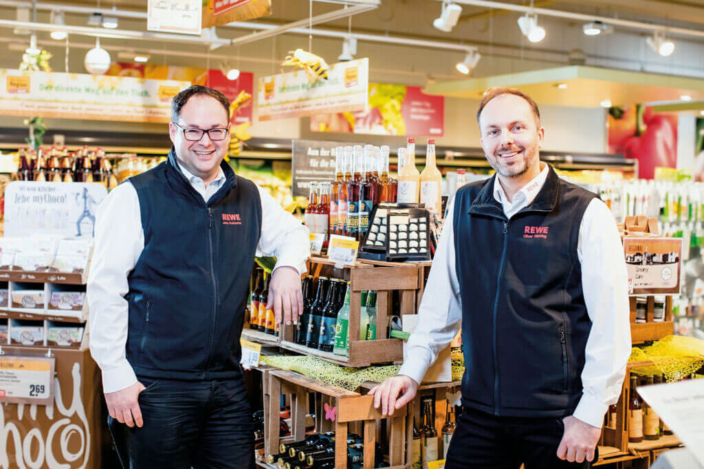 Jörg Kaluscha und Oliver Wehling in REWE-Dienstkleidung stehen lächelnd vor einem Verkaufsregal mit regionalen Produkten.