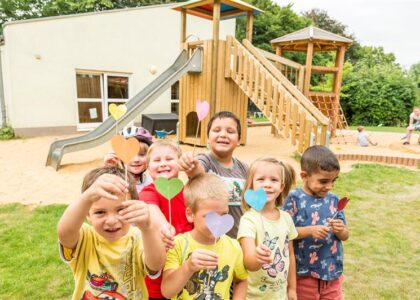 Fröhliche Kinder halten bunt gebastelte Papierherzen auf Holzstielen in die Kamera auf einem Spielplatz vor einem Klettergerüst.