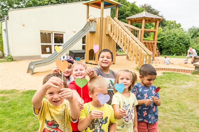 Fröhliche Kinder halten bunt gebastelte Papierherzen auf Holzstielen in die Kamera auf einem Spielplatz vor einem Klettergerüst.