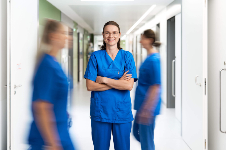 Lächelnde Pflegekraft in blauer Arbeitskleidung steht mit verschränkten Armen in einem hellen Krankenhausflur.
