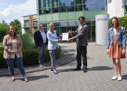 Fünf Personen stehen vor dem Klinikum Osnabrück, eine Frau und ein Mann überreichen sich gemeinsam eine gerahmte Urkunde.