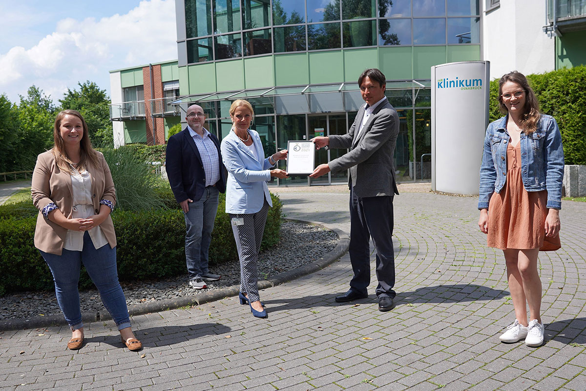 Fünf Personen stehen vor dem Klinikum Osnabrück, eine Frau und ein Mann überreichen sich gemeinsam eine gerahmte Urkunde.