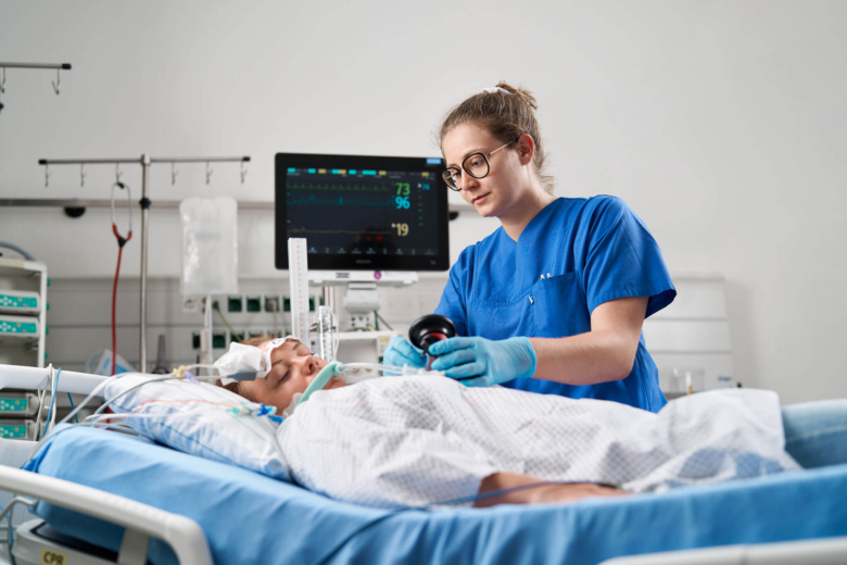Pflegekraft in blauer Kleidung bedient ein medizinisches Gerät bei einer liegenden Patientin im Intensivbett.