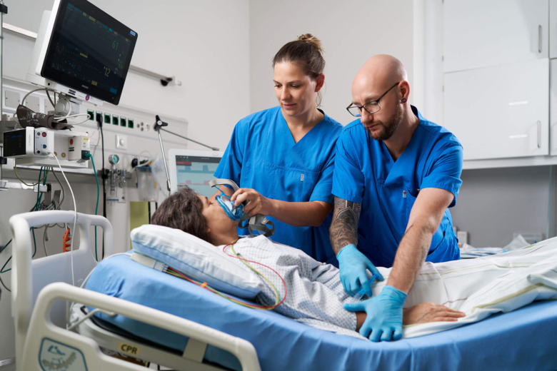 Zwei Pflegekräfte in blauer Arbeitskleidung versorgen gemeinsam eine Patientin im Krankenbett mit einer Atemmaske.