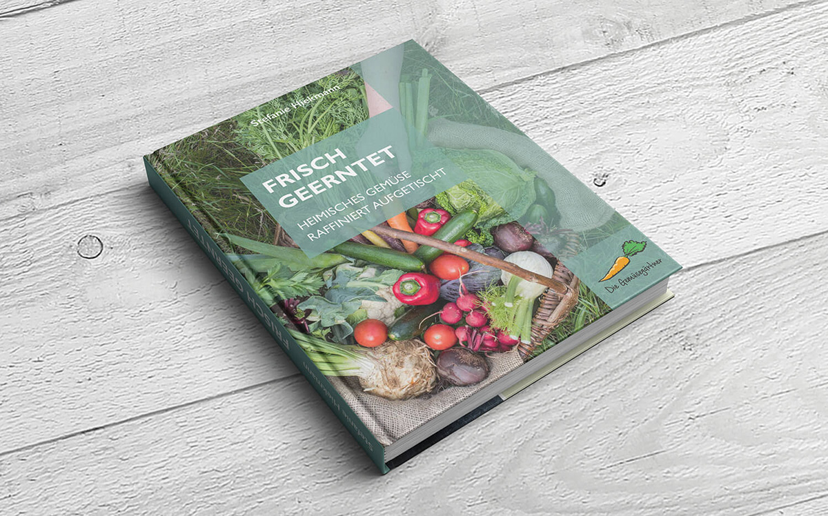 Das Kochbuch „Frisch geerntet“ mit einem bunten Gemüse-Cover liegt schräg auf einer hellen Holzfläche.