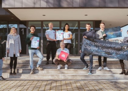 Eine Gruppe von Menschen steht vor einem Gebäude, präsentiert Urkunden sowie ein Banner des Kompetenzzentrums Digitale Transformation.