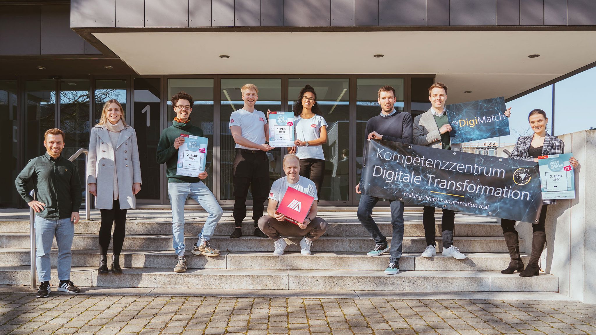 Eine Gruppe von Menschen steht vor einem Gebäude, präsentiert Urkunden sowie ein Banner des Kompetenzzentrums Digitale Transformation.