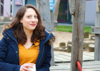 Kora Blanken im dunkelblauen Parka und gelben Oberteil sitzt mit einem Becher in der Hand auf einem Holzspielplatz.