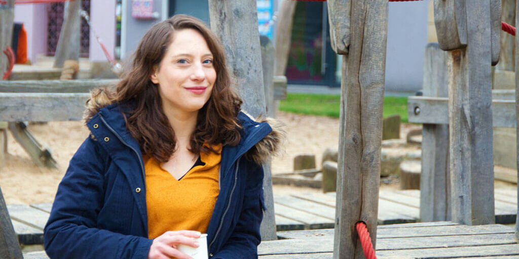 Kora Blanken im dunkelblauen Parka und gelben Oberteil sitzt mit einem Becher in der Hand auf einem Holzspielplatz.
