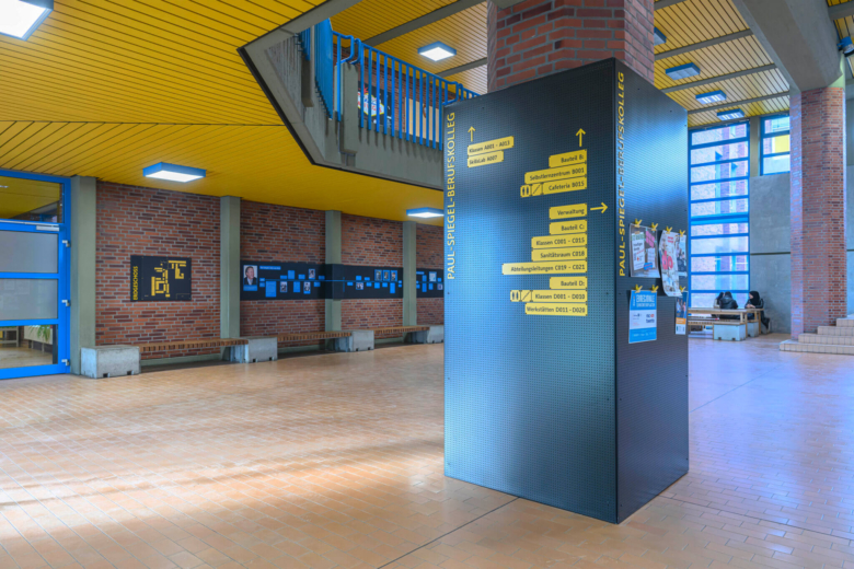Große schwarze Orientierungs-Stele mit gelber Beschriftung im Foyer des Paul-Spiegel-Berufskollegs.