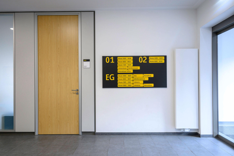 Schwarze Orientierungstafel mit gelber Schrift an einer weißen Wand neben einer Holztür.