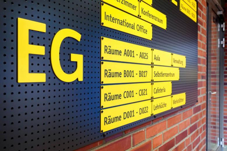 Nahaufnahme einer schwarzen Lochblechplatte an einer Klinkerwand mit gelben Wegweiser-Schildern für das Erdgeschoss (EG).