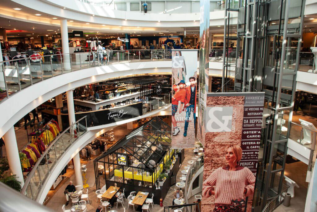 Innenansicht eines mehrstöckigen Kaufhauses (L&T). Man blickt von oben in das Atrium mit Rolltreppen, verschiedenen Geschäften und einem Café im Erdgeschoss. Große Werbebanner hängen von der Decke.