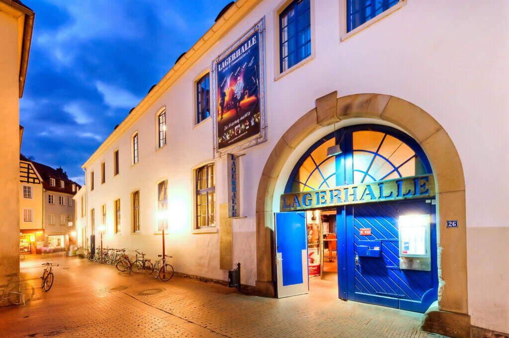 Außenansicht des Kulturzentrums „Lagerhalle“ bei Dämmerung mit beleuchtetem, großem blauen Eingangstor und Fahrrädern an der Hauswand.