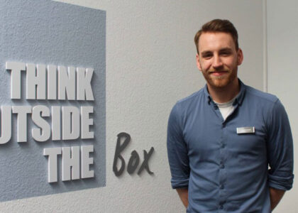 Portraitfoto von Lars Ruelander, lächelnd, neben einem Wandtattoo mit dem Schriftzug "THINK OUTSIDE THE BOX".