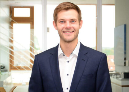Porträt von Lennard Koch im dunkelblauen Anzug und weißem Hemd vor einem hellen Bürohintergrund mit Glaswänden.