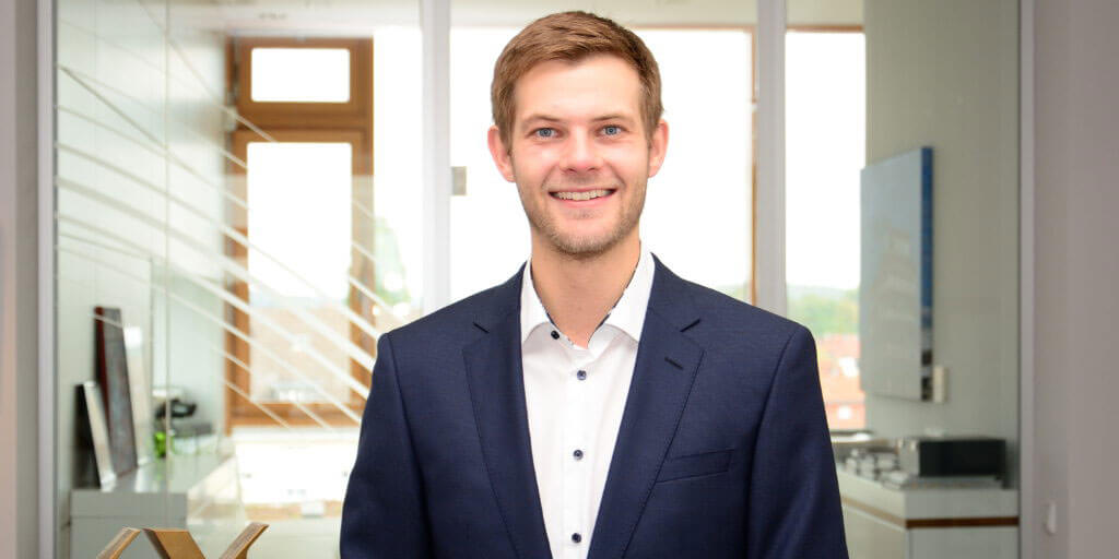 Porträt von Lennard Koch im dunkelblauen Anzug und weißem Hemd vor einem hellen Bürohintergrund mit Glaswänden.