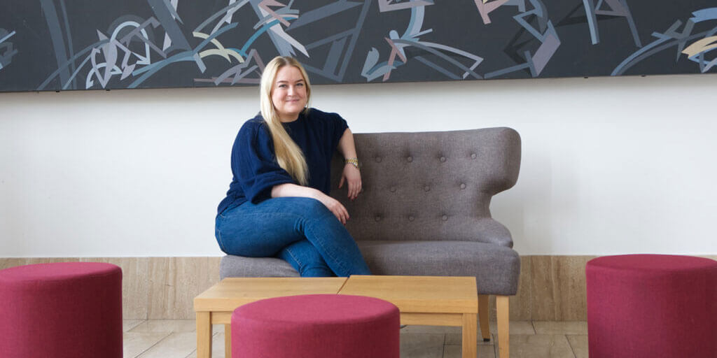 Leonie Ludewig sitzt lächelnd auf einem grauen Sofa vor einer Wand mit moderner Kunst.