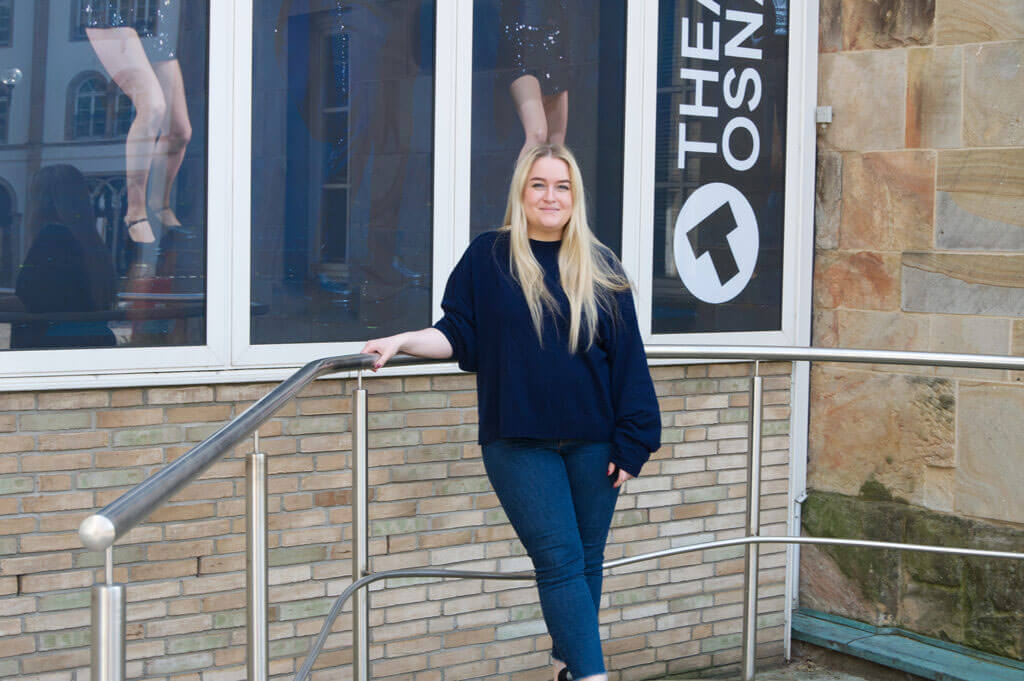 Leonie Ludewig steht lächelnd an einem Metallgeländer vor dem Gebäude des Theaters Osnabrück.