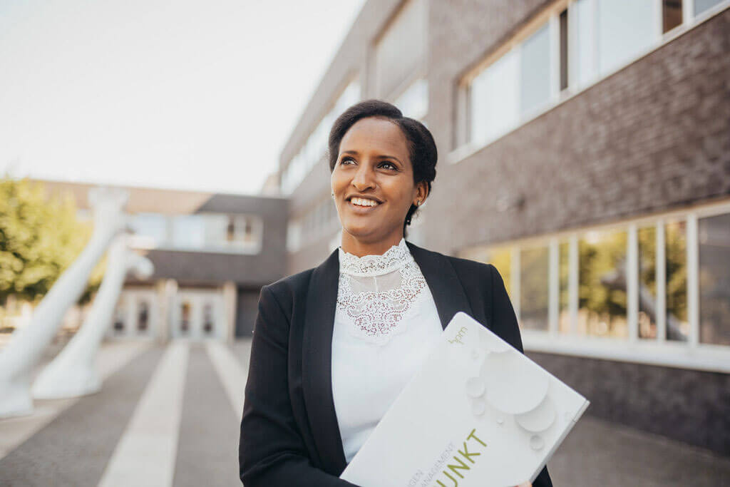 Lächelnde Frau im schwarzen Blazer blickt zuversichtlich nach oben und hält eine Infobroschüre der Universität Osnabrück im Arm.