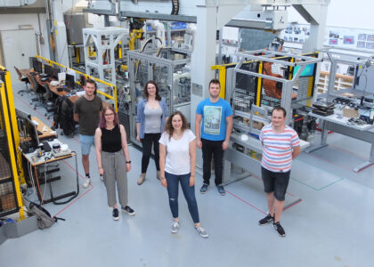 Sechs junge Menschen stehen lächelnd in einer modernen Industriehalle mit Roboteranlagen und Computerarbeitsplätzen.