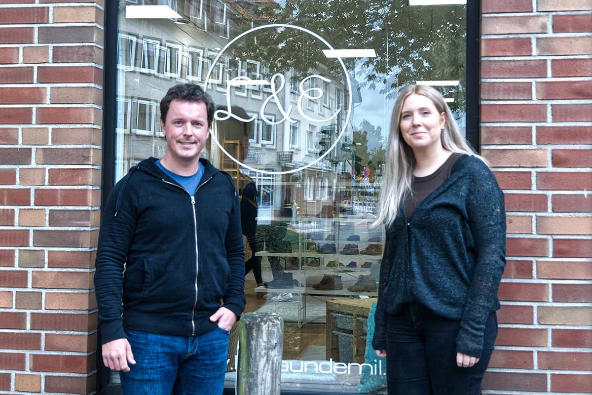 Ein Mann und eine Frau stehen lächelnd vor dem Schaufenster eines Geschäfts mit dem Logo „L&E“ auf der Scheibe.