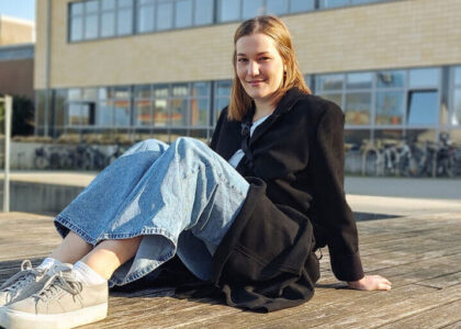 Louisa Peters sitzt lächelnd in lässiger Kleidung auf einem Holzdeck im Freien vor einem modernen Universitätsgebäude.