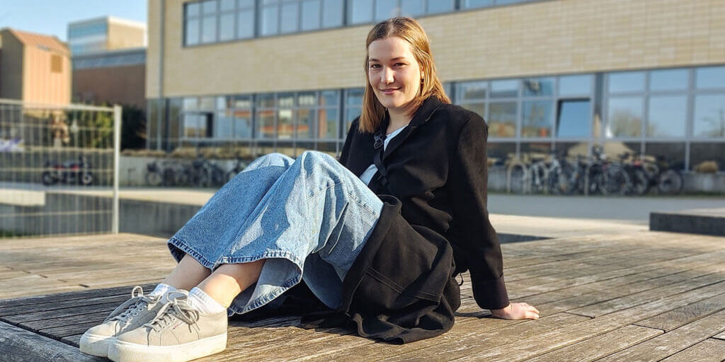 Louisa Peters sitzt lächelnd in lässiger Kleidung auf einem Holzdeck im Freien vor einem modernen Universitätsgebäude.