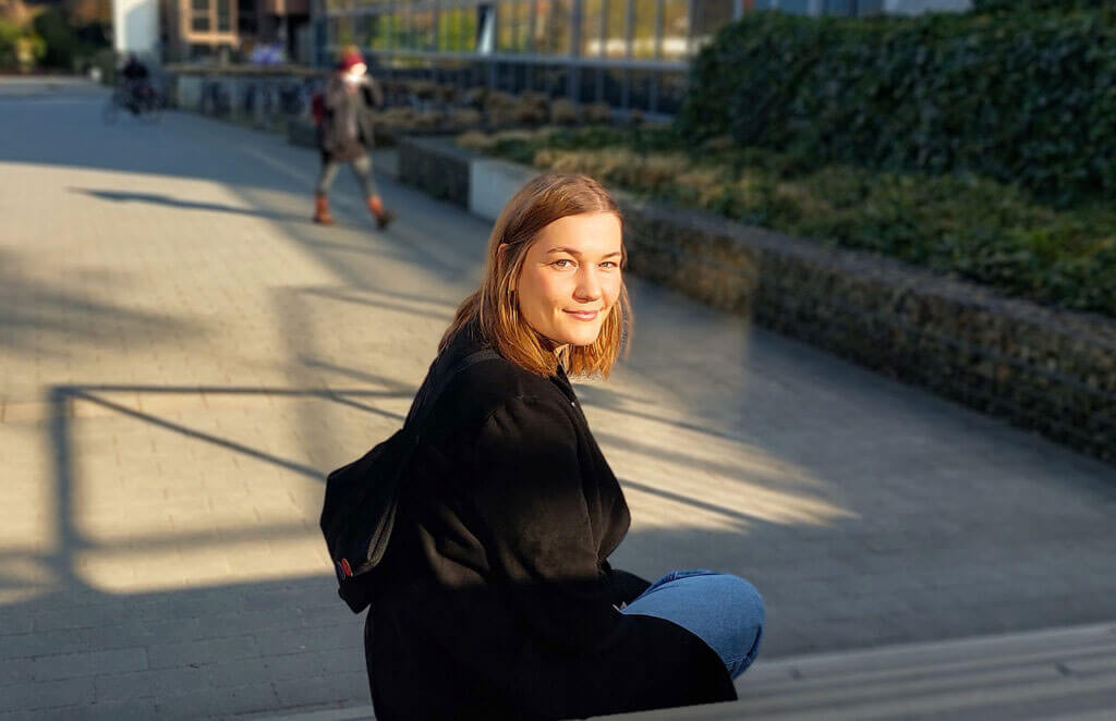 Porträt von Louisa Peters, die im Gegenlicht auf einer Außentreppe sitzt und lächelnd zurückblickt.