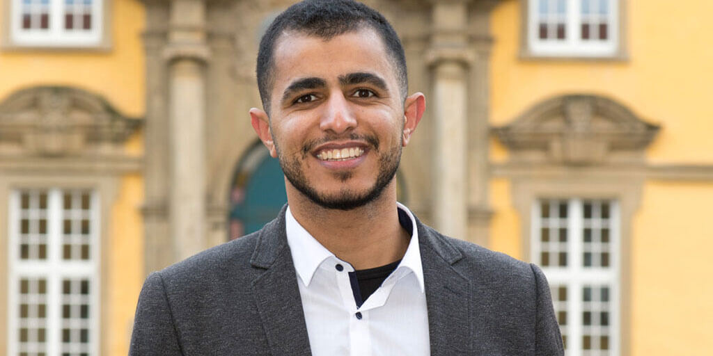 Portrait von Mahmoud Farrag im grauen Sakko und weißen Hemd vor einem historischen, gelben Gebäude.