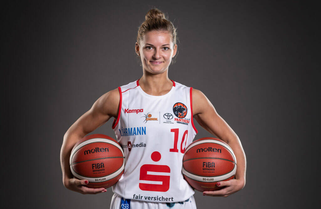 Mannschaftskapitänin Jenny Strozyk steht im weiß-roten Trikot vor grauem Hintergrund und hält lächelnd zwei Basketbälle im Arm.