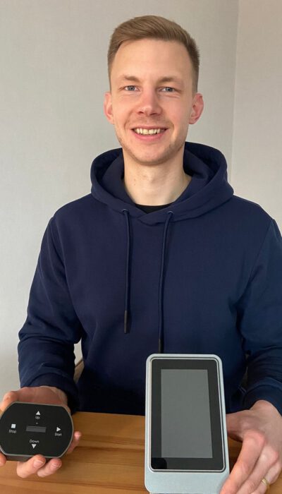 Ein lächelnder Mann in blauem Hoodie zeigt eine Fernbedienung und ein Tablet mit schwarzem Bildschirm.