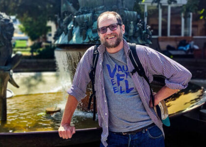 Matthew Rees steht mit Brille, Rucksack und offenem Hemd lächelnd vor einem großen Springbrunnen im Freien.