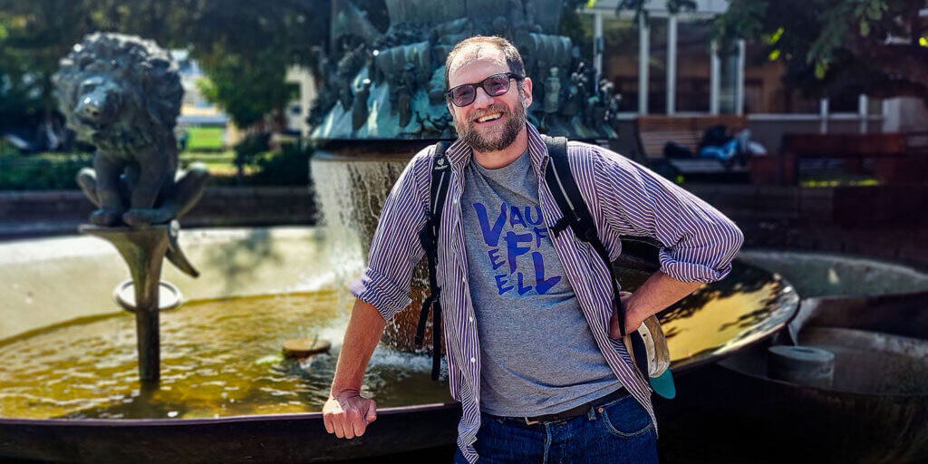 Matthew Rees steht mit Brille, Rucksack und offenem Hemd lächelnd vor einem großen Springbrunnen im Freien.
