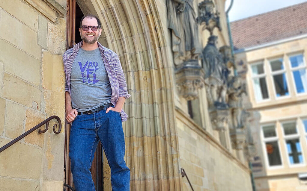 Matthew Rees lehnt sich lächelnd an den steinigen Torbogen eines historischen Rathauses in Osnabrück.